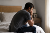Hombre real sentado de perfil en el borde de la cama con las manos en la cara, reflejando preocupación y el impacto emocional de la ansiedad de ejecución.